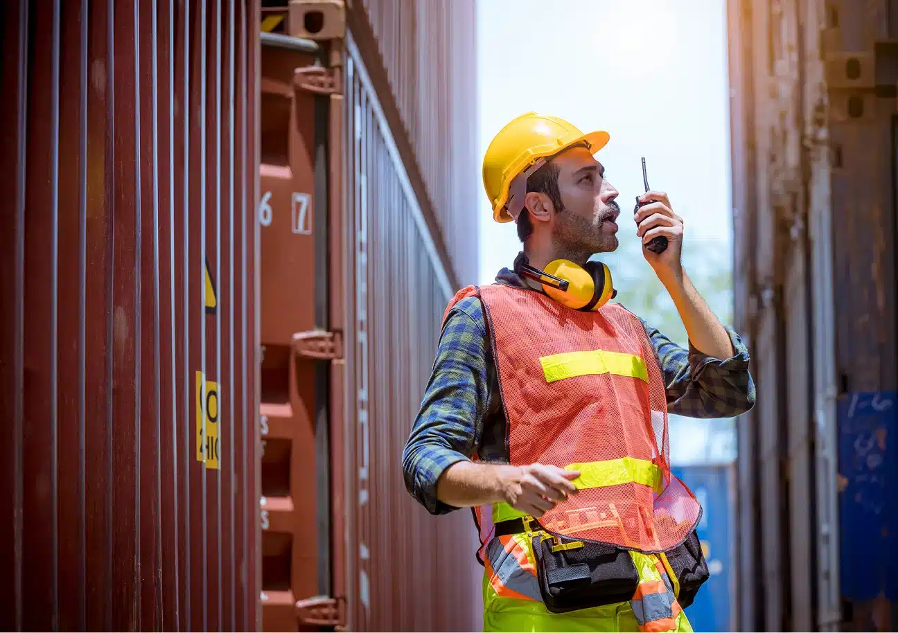 Worker in safety gear using walkie-talkie between shipping containers