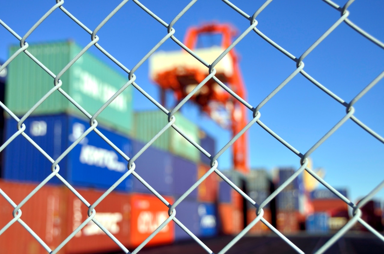 Chain-link fence with blurred shipping containers and crane in the background