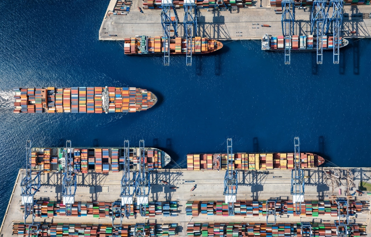 Aerial view of cargo ships docked at a busy port with numerous shipping containers and cranes.