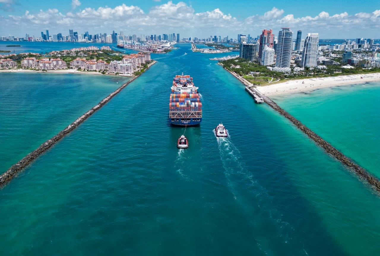 A large cargo ship with containers entering a port channel guided by two tugboats with city skyline in the background