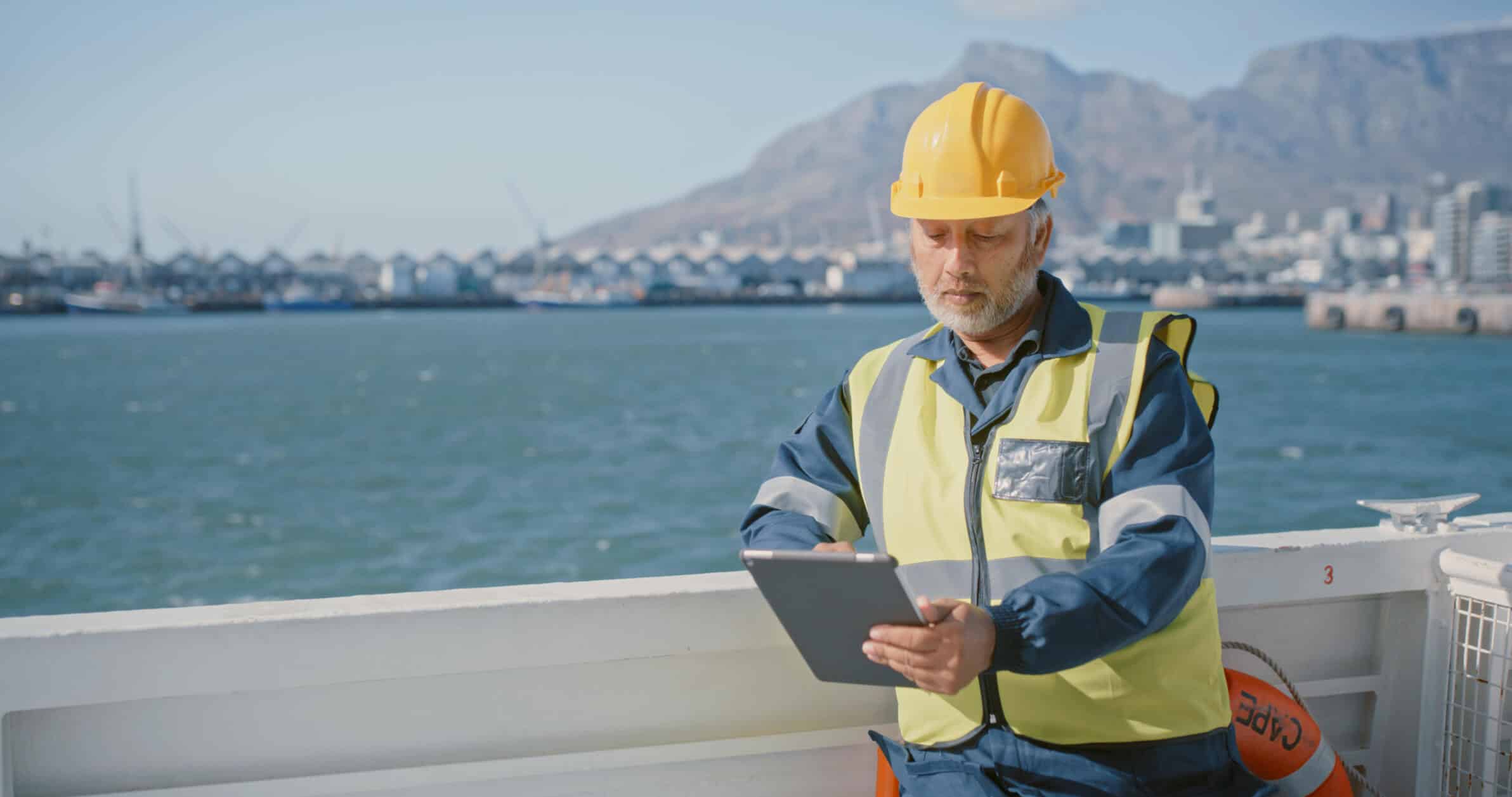Man wearing safety gear using a tablet on a vessel with harbor and mountains in background