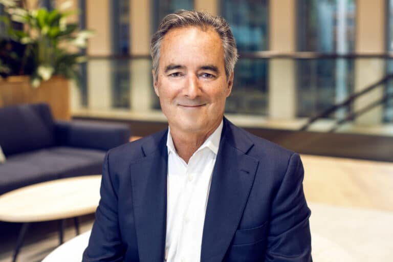 Man in a navy suit and white shirt sitting indoors with a slight smile