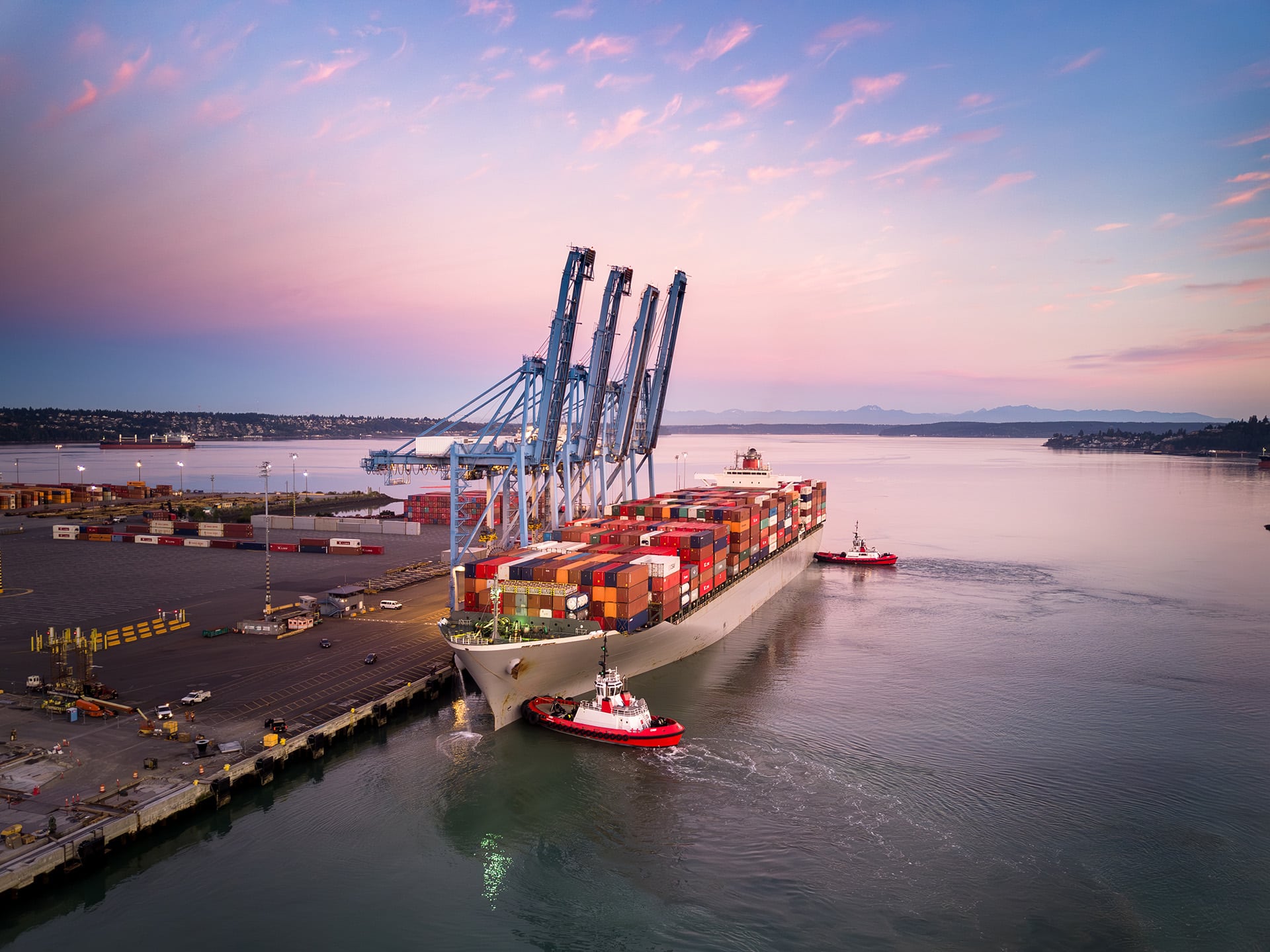 Cargo ship docked at port with cranes and tugboats during sunset